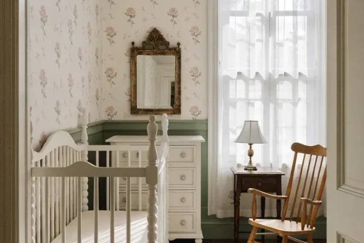 A vintage nursery with delicate pink rose wallpaper, an antique white crib, and soft cream bedding.