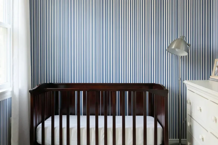 A traditional nursery with thin blue and white ticking stripe wallpaper and a dark wood crib.