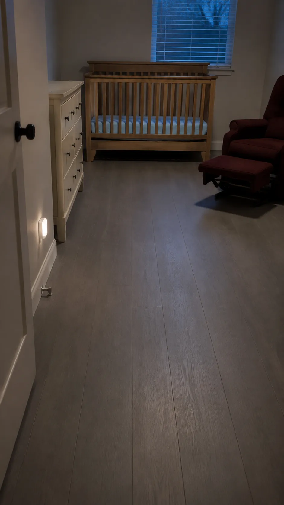 Photograph the clear walking route from a nursery door to the crib as the central subject, with the dresser and glider positioned outside that path