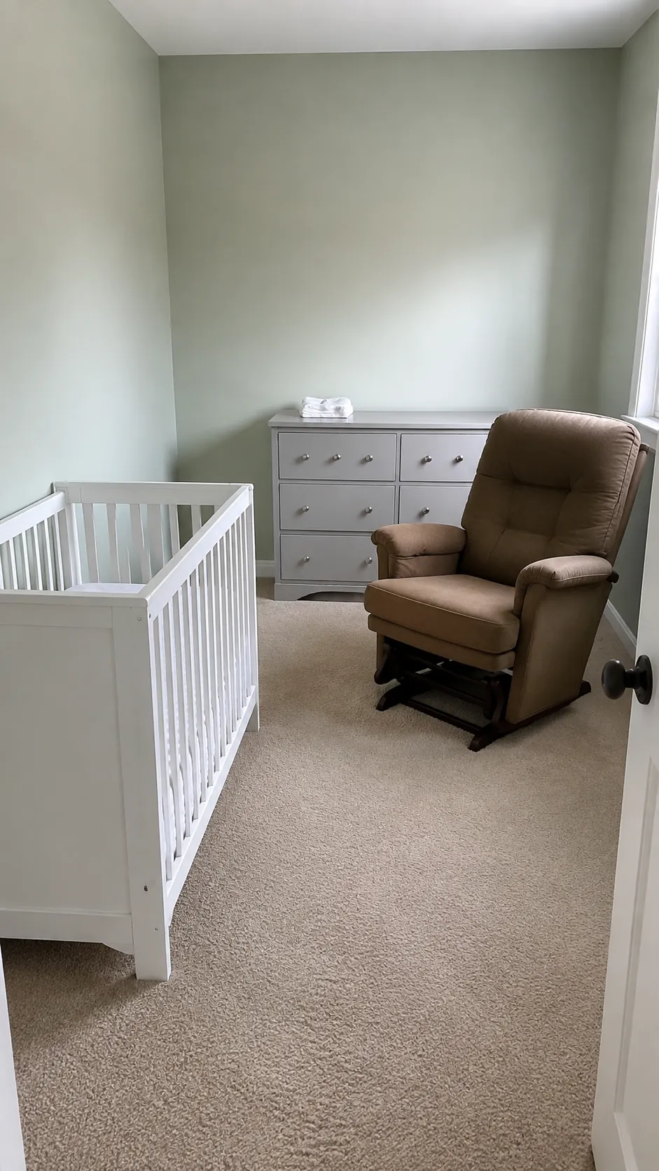 Show a compact mini crib as a deliberate furniture size choice that leaves enough usable space for a glider, rather than depicting a cramped full size cot