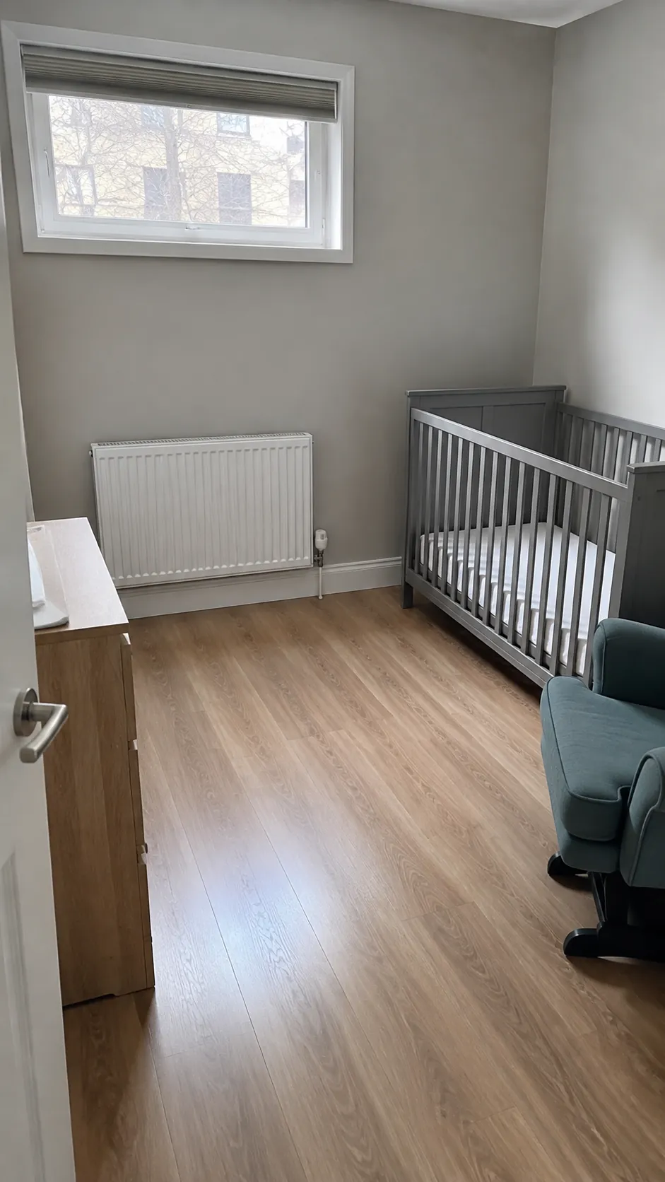 Illustrate a nursery that leaves a radiator wall genuinely clear while placing the crib, dresser, and glider elsewhere